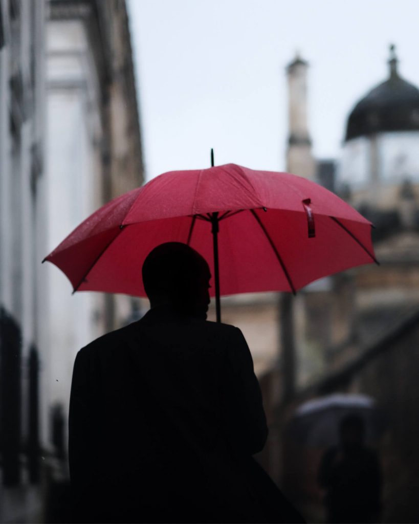 Back of a silhouetted man in a silhouetted city holding a red umbrella over his head