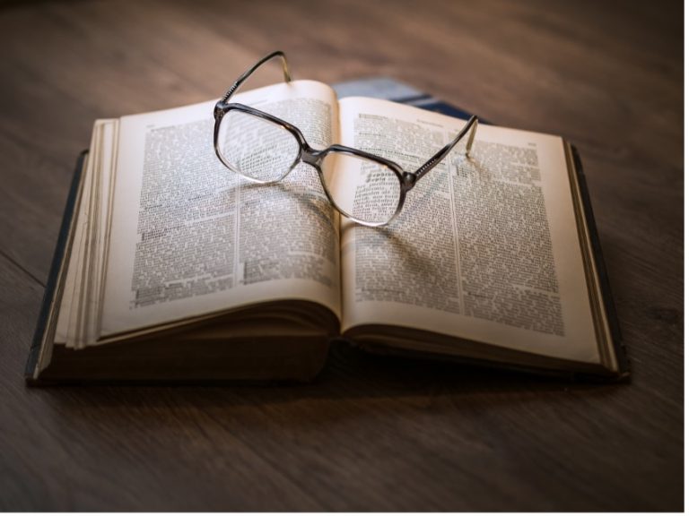 Alt text: An open book with small text, accompanied by a pair of glasses, representing the JROC Report's summary on Open Banking, Payment Initiation, and Fintech's Future.